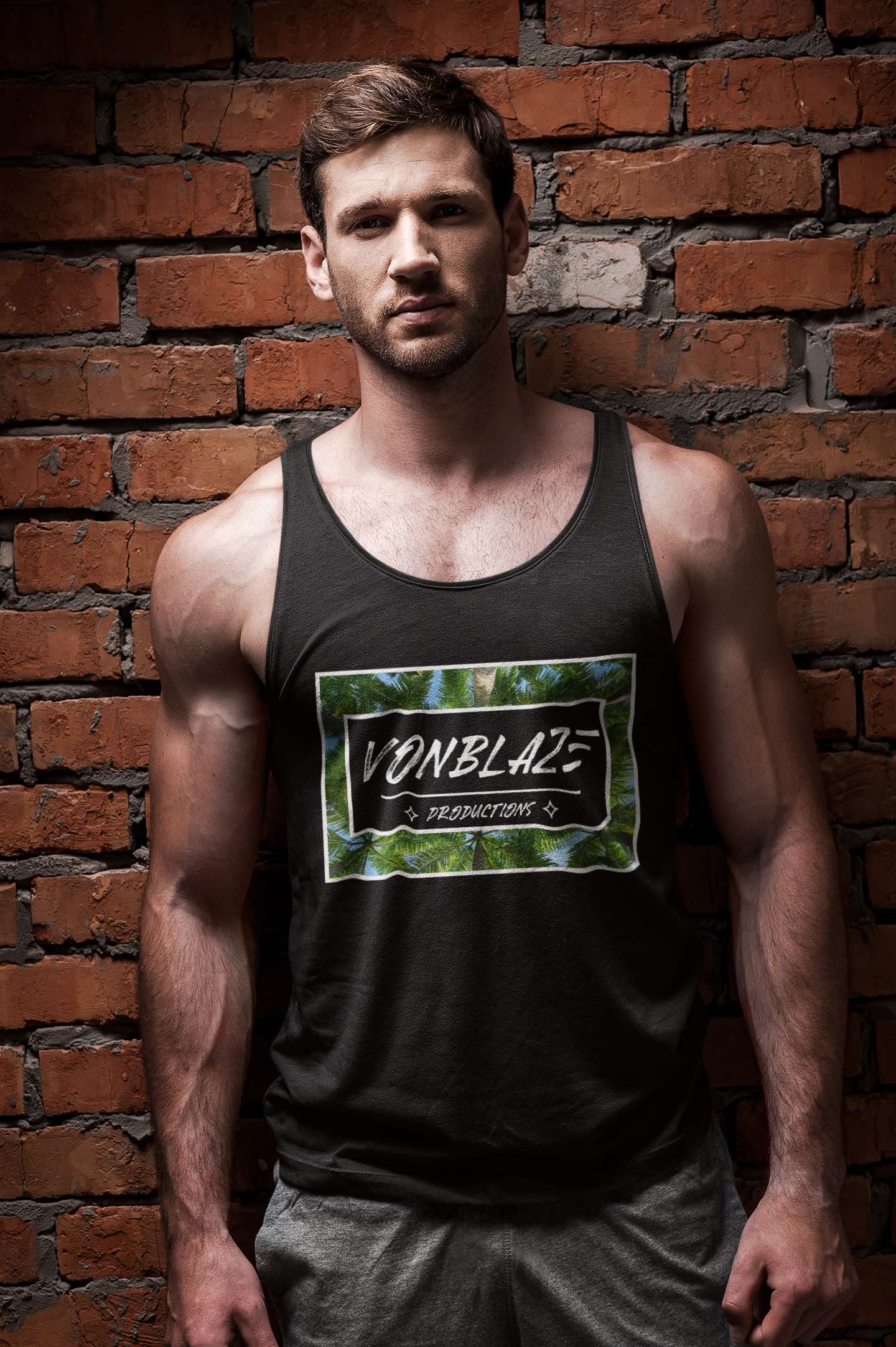 Muscular man standing infront of a brick wall wearing a black tank top featuring a logo with VonBlaze Productions in writing inside a black rectangle set on a palm tree backdrop