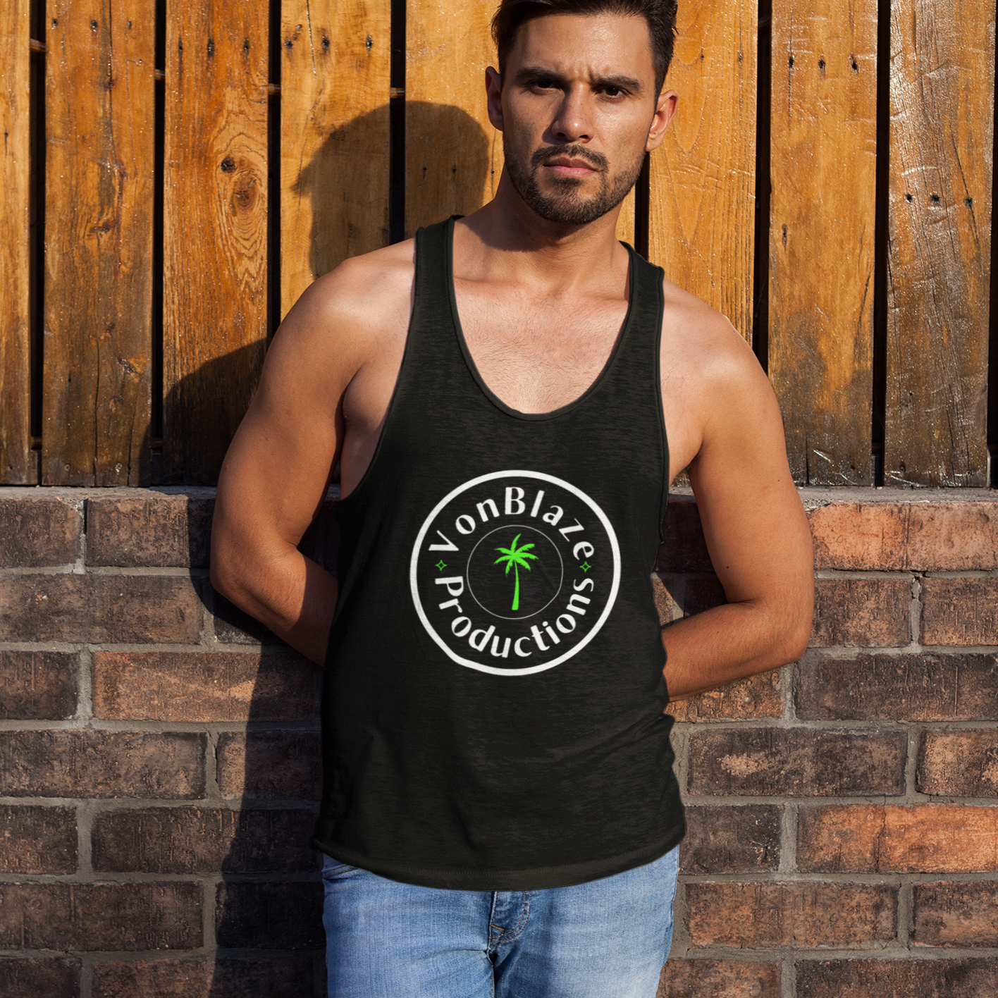 Muscular man leaning up against a brick wall and fence wearing a black tank top featuring a circular logo with the words VonBlaze Productions and a palm tree in the middle