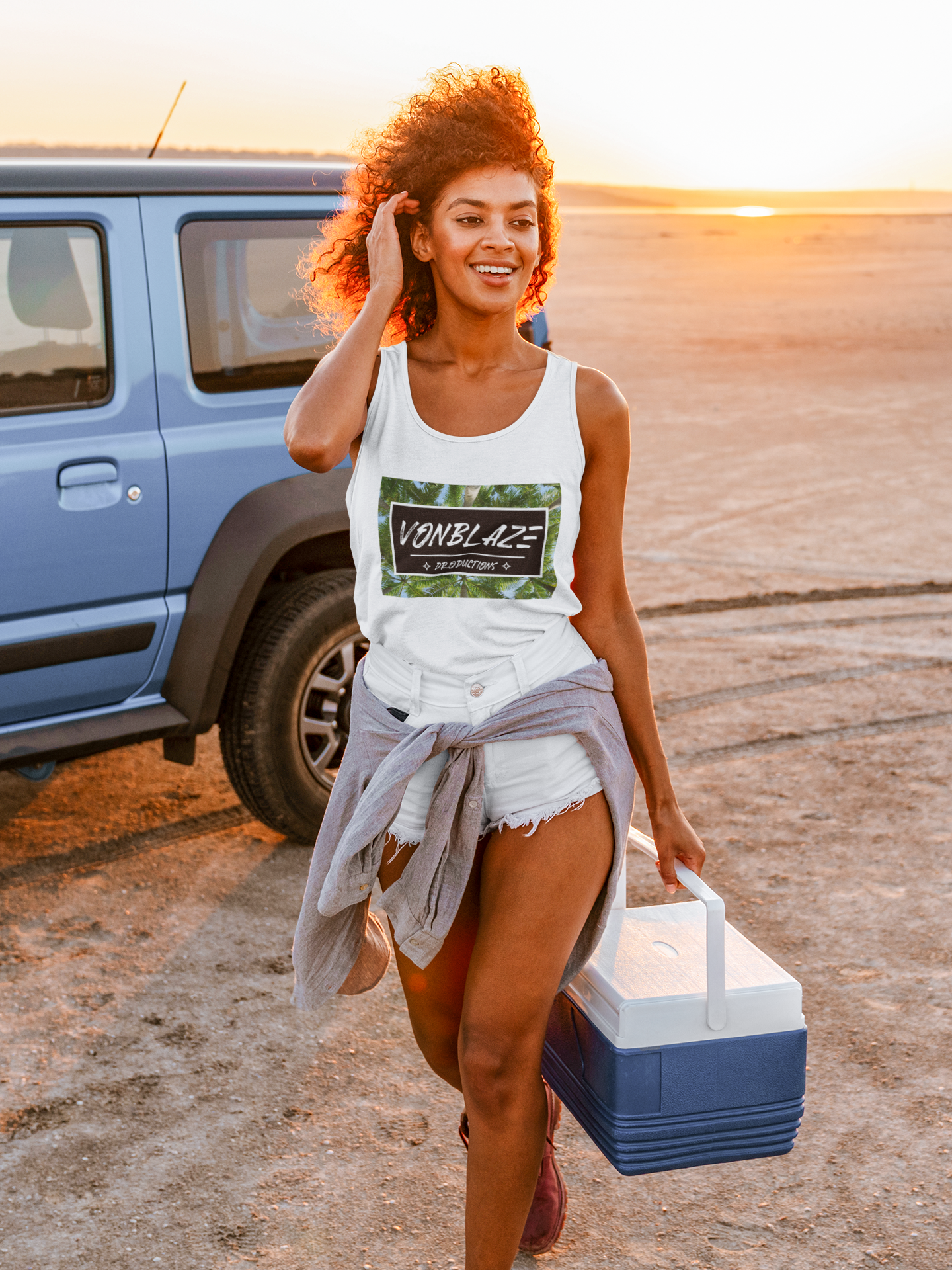 Young woman carrying a cooler on the beach wearing a white tank top featuring a logo with VonBlaze Productions in writing inside a black rectangle set on a palm tree backdrop