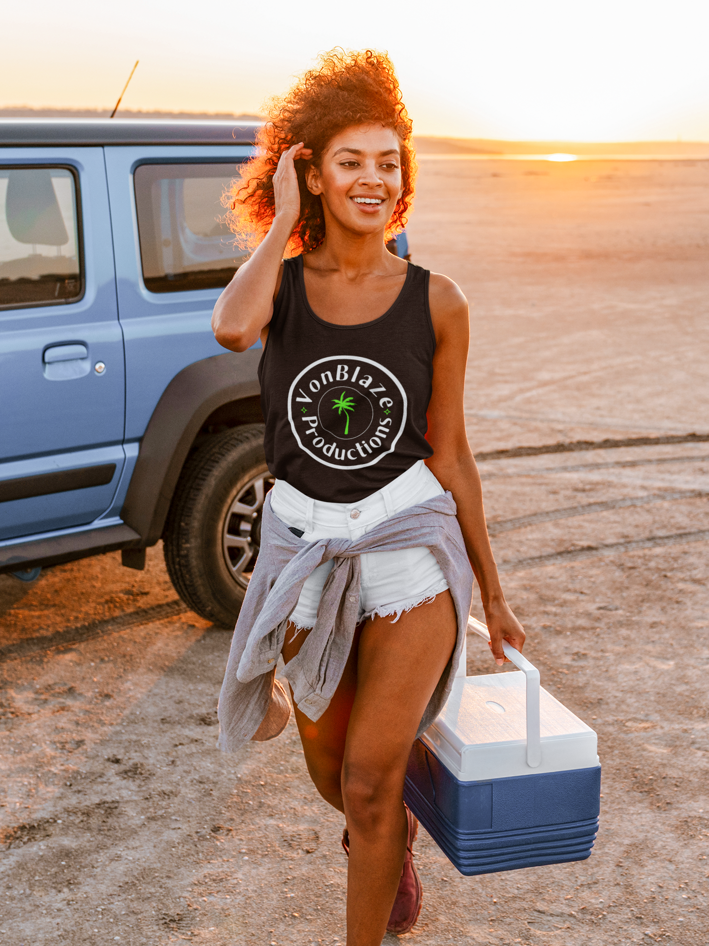 Young woman carrying a cooler on the beach wearing a black tank top featuring a circular logo with the words VonBlaze Productions and a palm tree in the middle