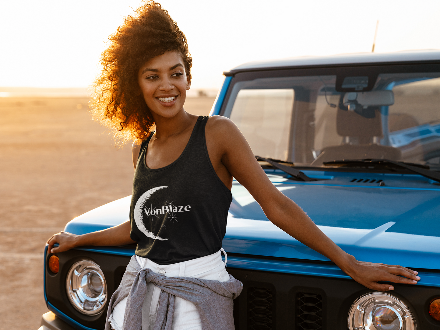 Young woman leaning up against the hood of a truck on the beach while wearing a black tank top featuring a logo that says VonBlaze Productions with a half crescent moon and stars